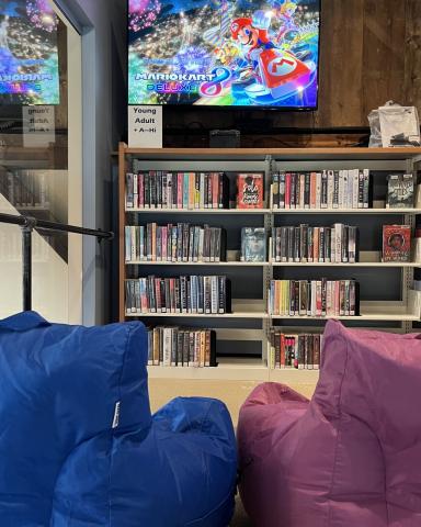 Image of TV displaying Mario Kart and one blue with one purple bean bag chair. Book shelf along wall.